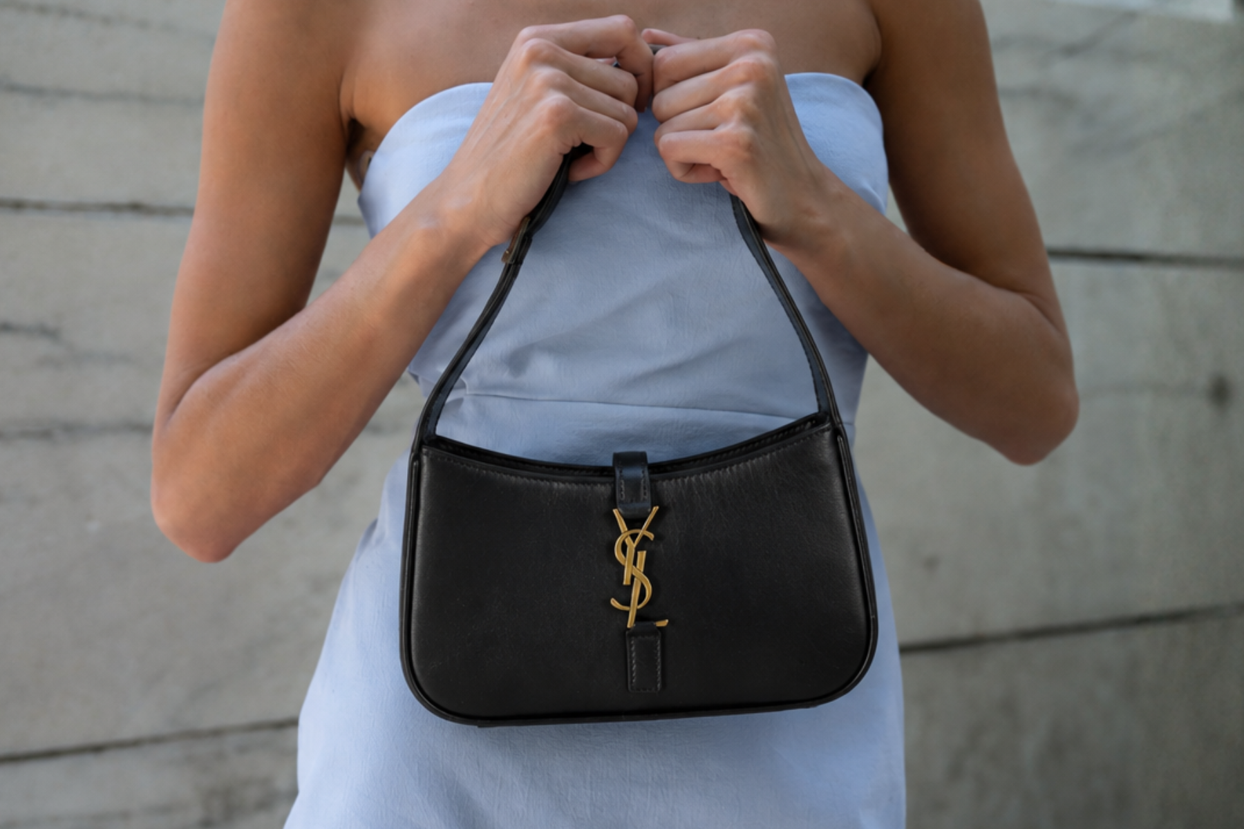 A black leather shoulder bag with a gold-tone YSL monogram clasp, held by a model wearing a light blue strapless dress against a gray concrete wall.