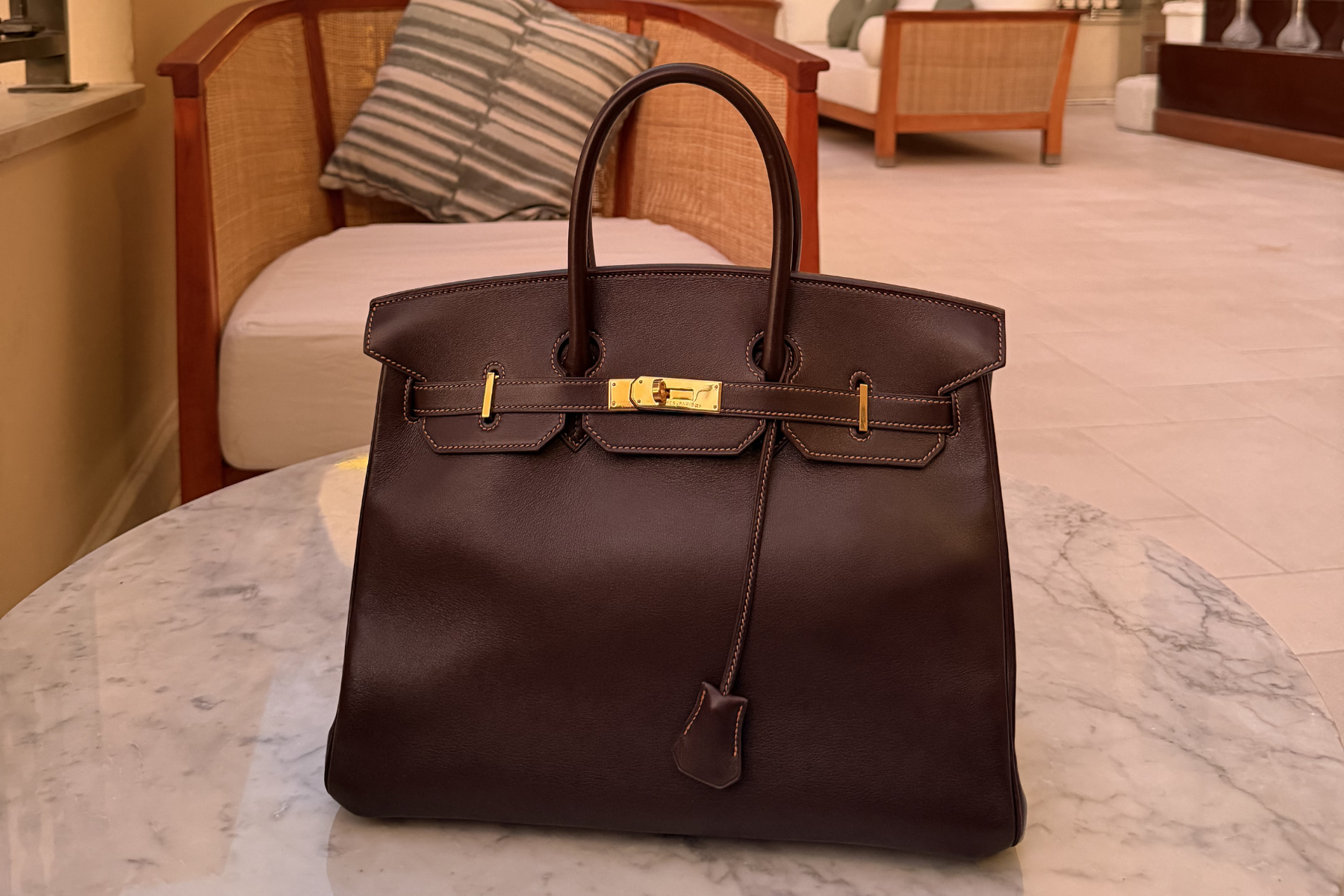 A dark brown leather Hermès Birkin handbag with gold-tone hardware, displayed on a white marble table in a modern living room setting.