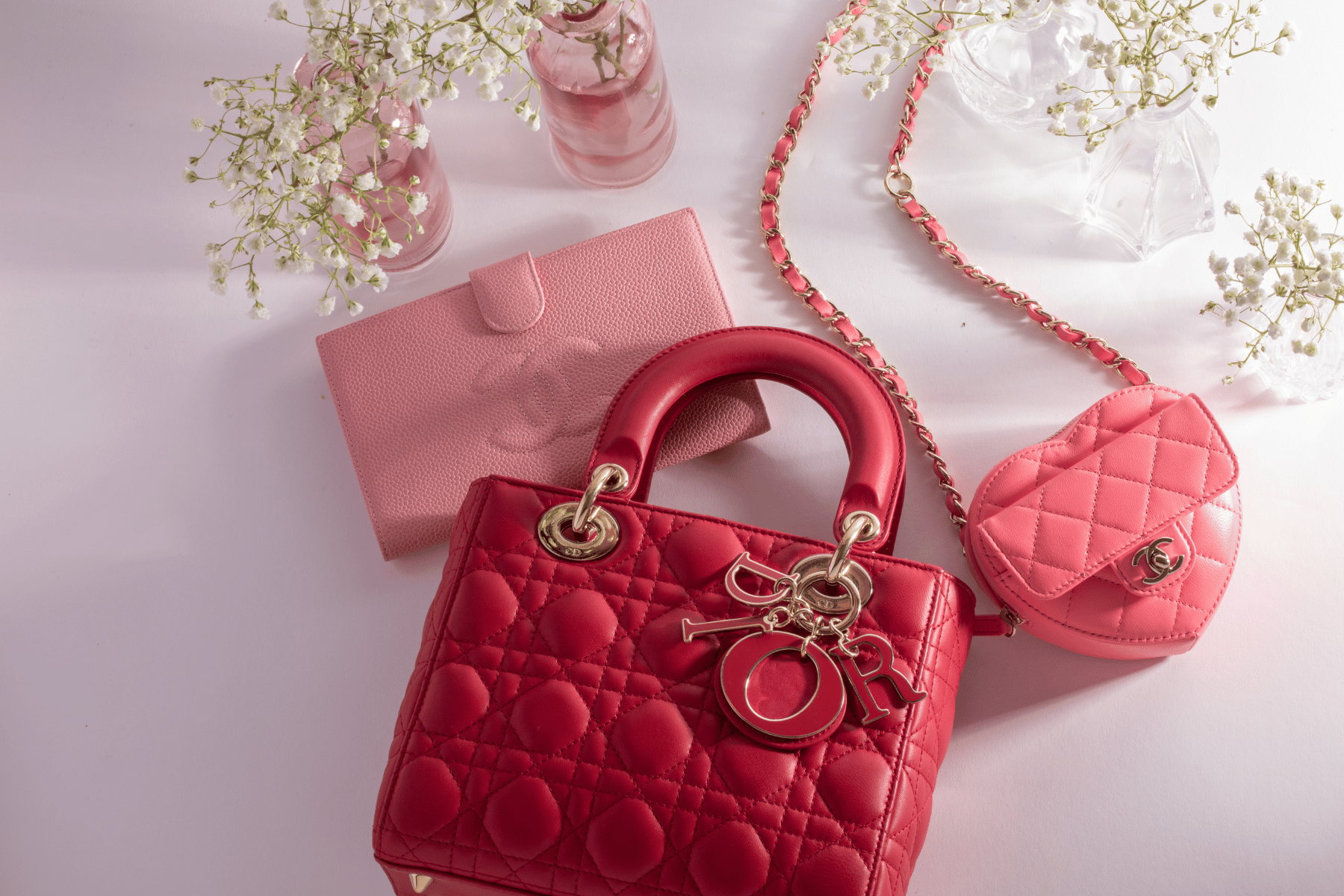 Red Dior handbag with gold accents and pink Chanel Heart bag and wallet on a white background with flowers