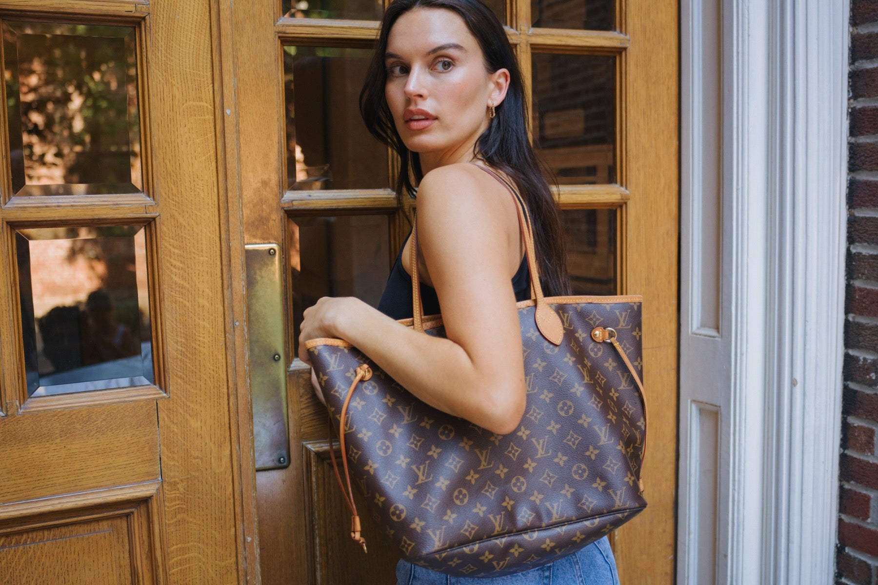 Woman holding a brown LV Neverfull monogram handbag in front of a wooden door.