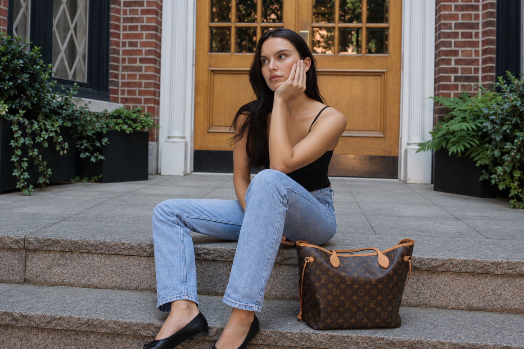 A woman sits on stone steps in front of a brick building, wearing a black camisole, light-wash blue jeans, and black flats. A brown Louis Vuitton monogram Neverfull bag rests beside her.